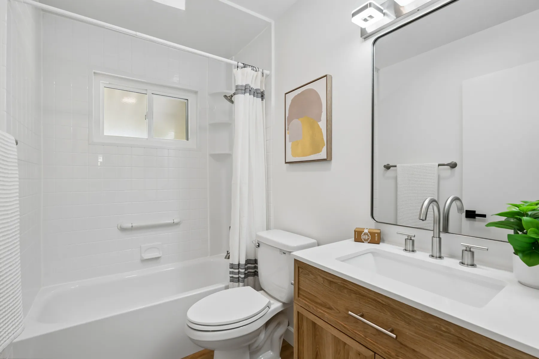Second bathroom with a wood-tone vanity, a white tub and shower surround, and a large black-framed mirror.